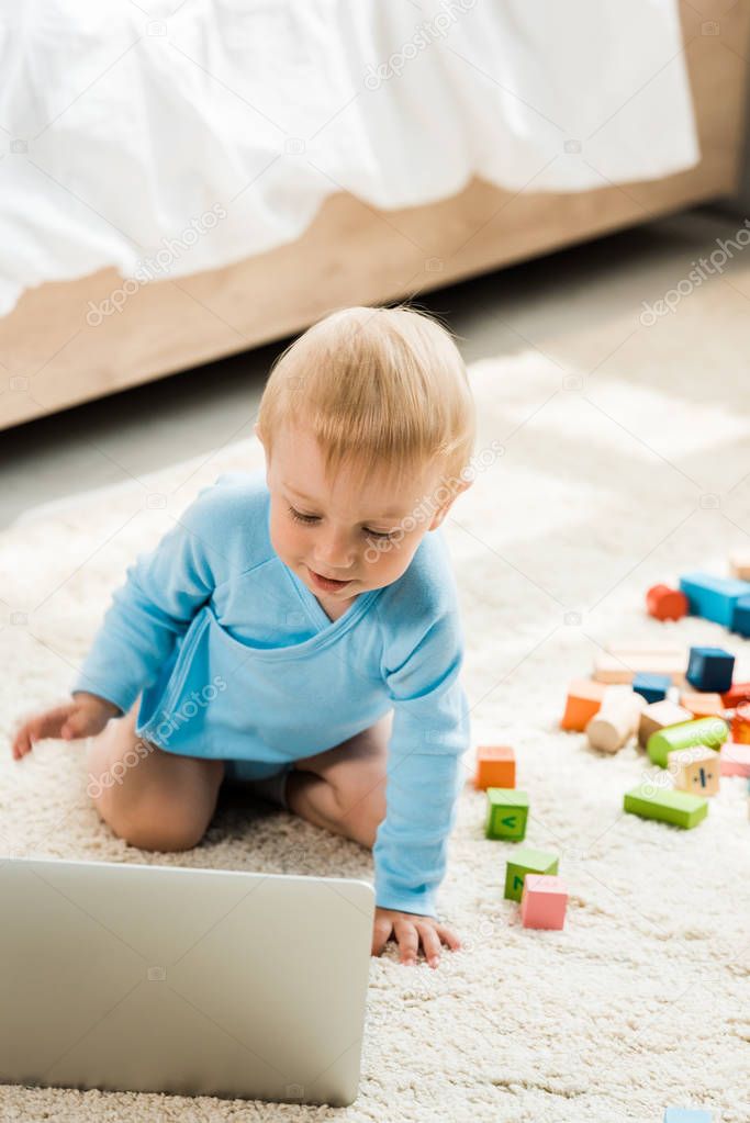 selective focus of happy toddler kid looking at laptop near colorful toy blocks on carpet 