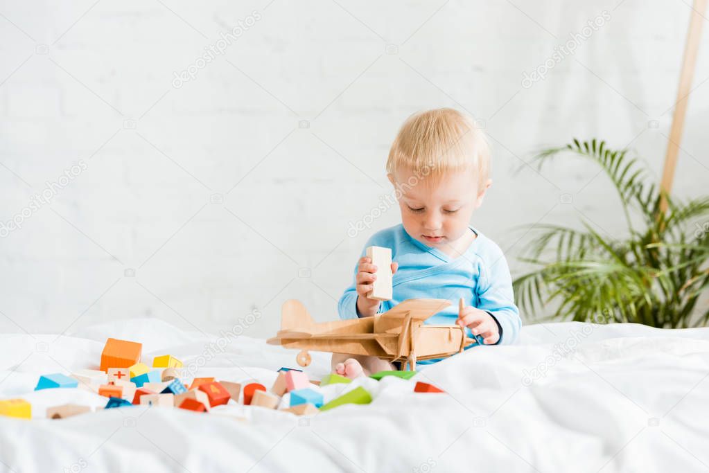 cute toddler boy playing with wooden biplane near colorful toy blocks on bed 