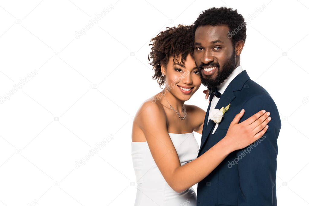 cheerful african american bridegroom and bride hugging and looking at camera isolated on white 