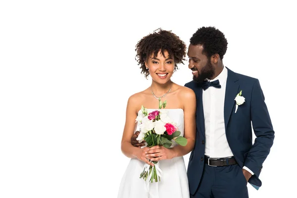 白で隔離されたポケットに手を入れて立っている花婿の近くに花束を持つ幸せなアフリカ系アメリカ人の花嫁 — ストック写真
