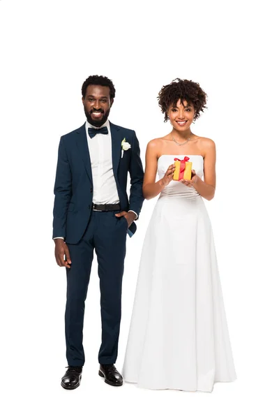 陽気な花嫁の近くにポケットに手を入れて立っている幸せなアフリカ系アメリカ人の花婿は 白に隔離されたギフトボックスを保持しています — ストック写真