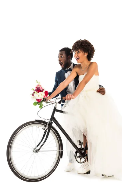 白に隔離された自転車に乗って花を持つ陽気な花嫁の近くに驚いたアフリカ系アメリカ人の花婿 — ストック写真