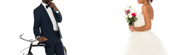 панорамный снимок африканского жениха, стоящего рядом с велосипедом, и счастливой невесты с цветами, изолированными на белом
 