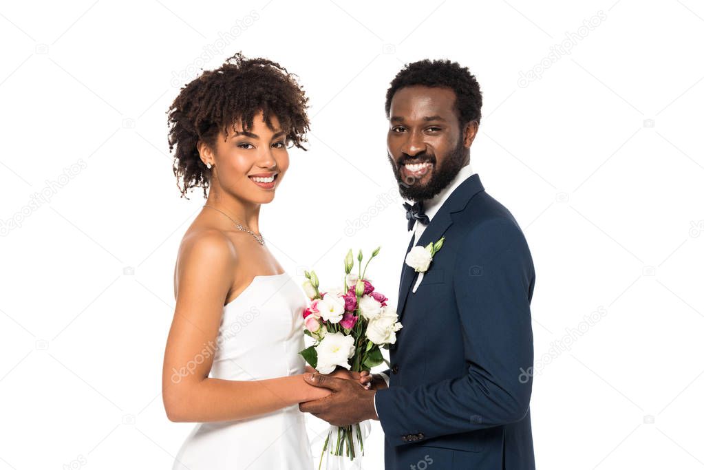 cheerful african american bride holding bouquet near bridegroom and looking at camera isolated on white 