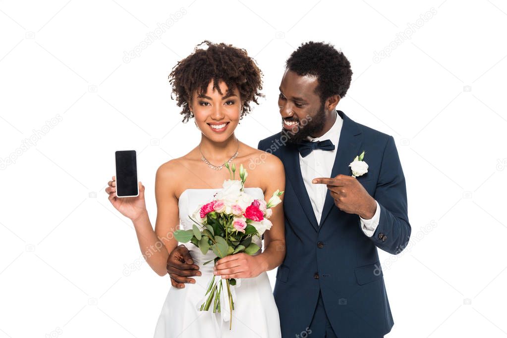 happy african american bridegroom pointing with finger at smartphone with blank screen near bride isolated on white 