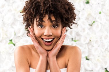 happy african american bride touching face while looking at camera near flowers  clipart