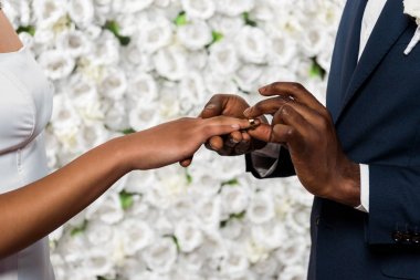 cropped view of african american man putting wedding ring on finger of bride near flowers  clipart