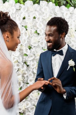 happy african american man putting wedding ring on finger of bride near flowers  clipart