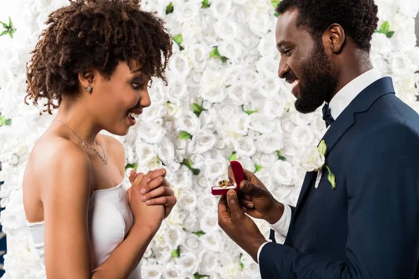 花の近くに陽気な花嫁の近くにリングとボックスを保持している幸せなアフリカ系アメリカ人の男性 — ストック写真