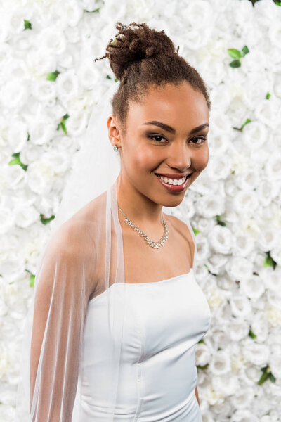 happy african american bride in white veil smiling near flowers 