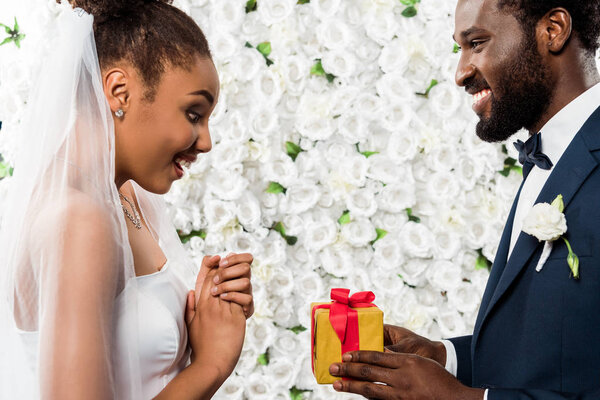 surprised african american bride looking at present near happy bridegroom and flowers 