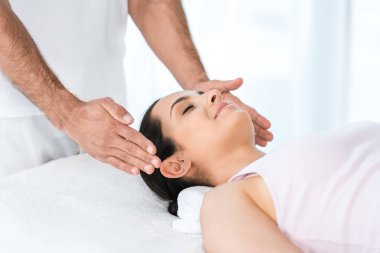 masaj masasında yatan kapalı gözleri ile mutlu kadının başının yakın ellerini koyarak şifacı kırpılmış görünümü