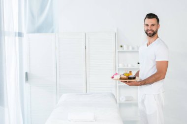 neşeli sakallı adam yağ şişeleri ile tepsi tutan, spa merkezinde deniz tuzu ve çiçekler ile kase 