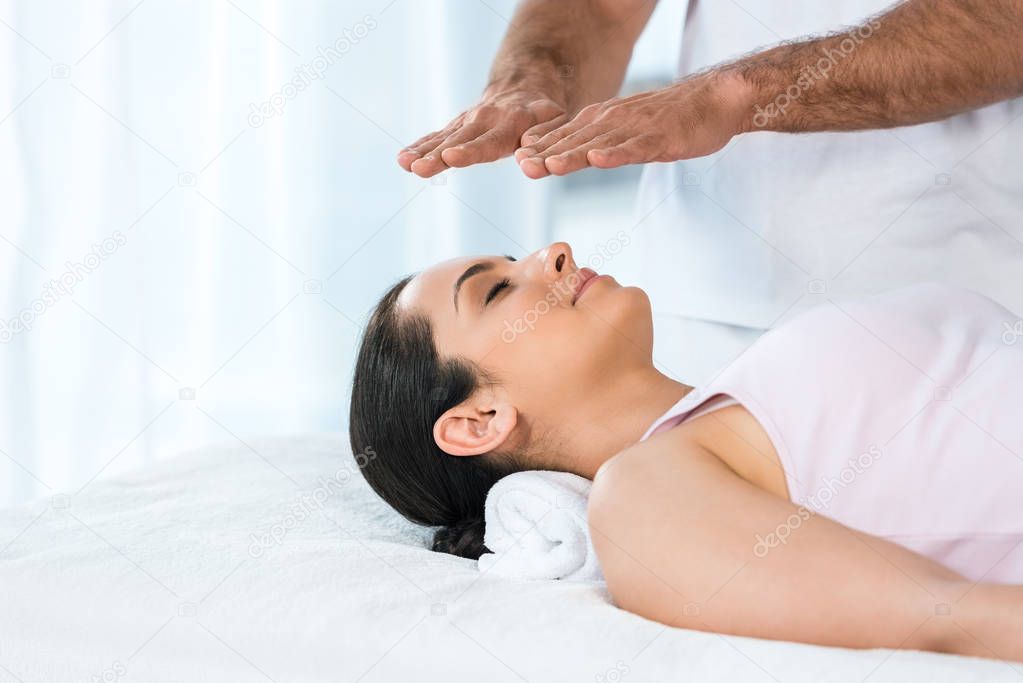 cropped view of man putting hands above head of attractive woman with closed eyes lying on massage table