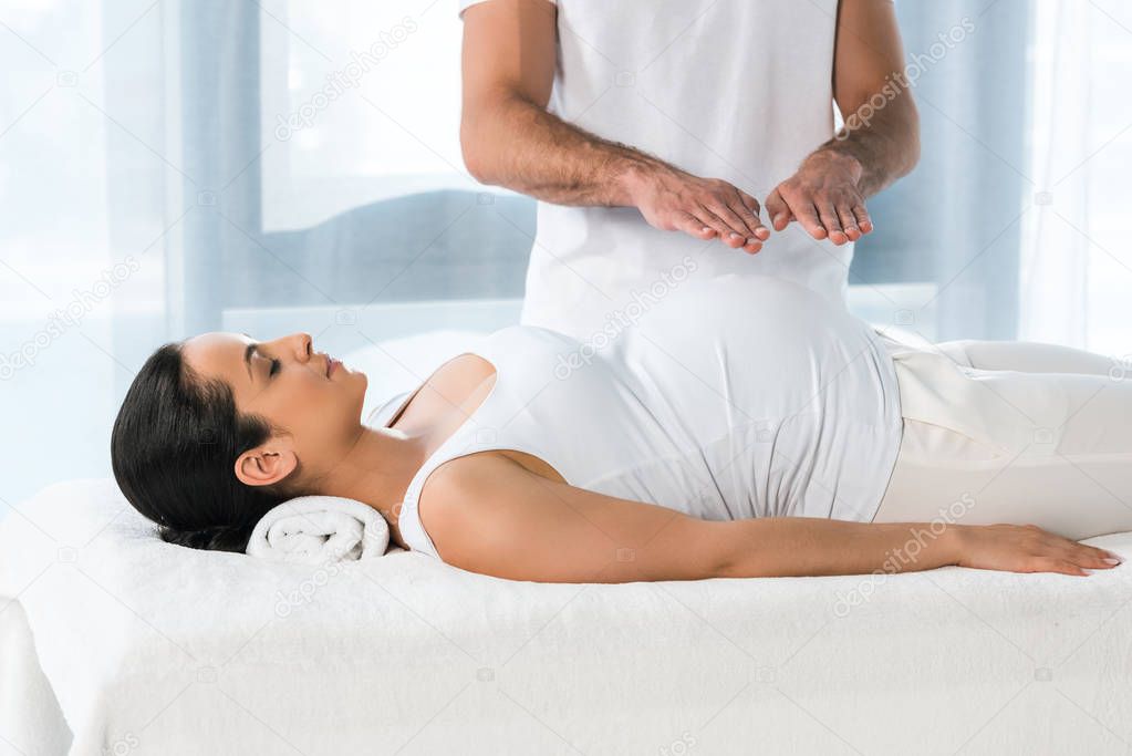 cropped view of healer putting hands above tummy of brunette pregnant woman 