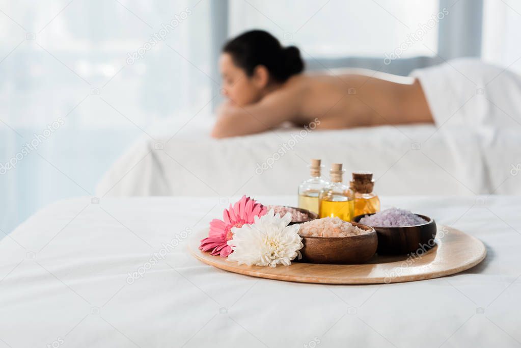 selective focus of tray with bottles of oil, bowls with sea salt and flowers near brunette woman in spa center 