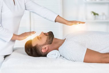 cropped view of healer putting hands above head while healing handsome man on massage table clipart