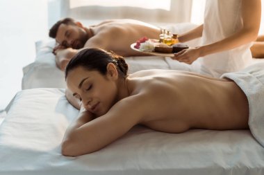 spa merkezinde kadın ve erkek yakın deniz tuzu ve yağ şişeleri ile tepsi tutan masör seçici odak 