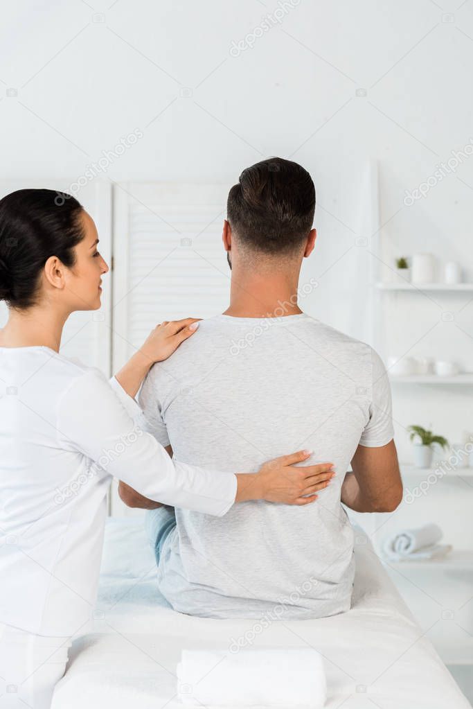 back view of man sitting on massage table near attractive healer 