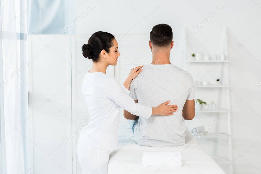 back view of man sitting on massage table near brunette healer 