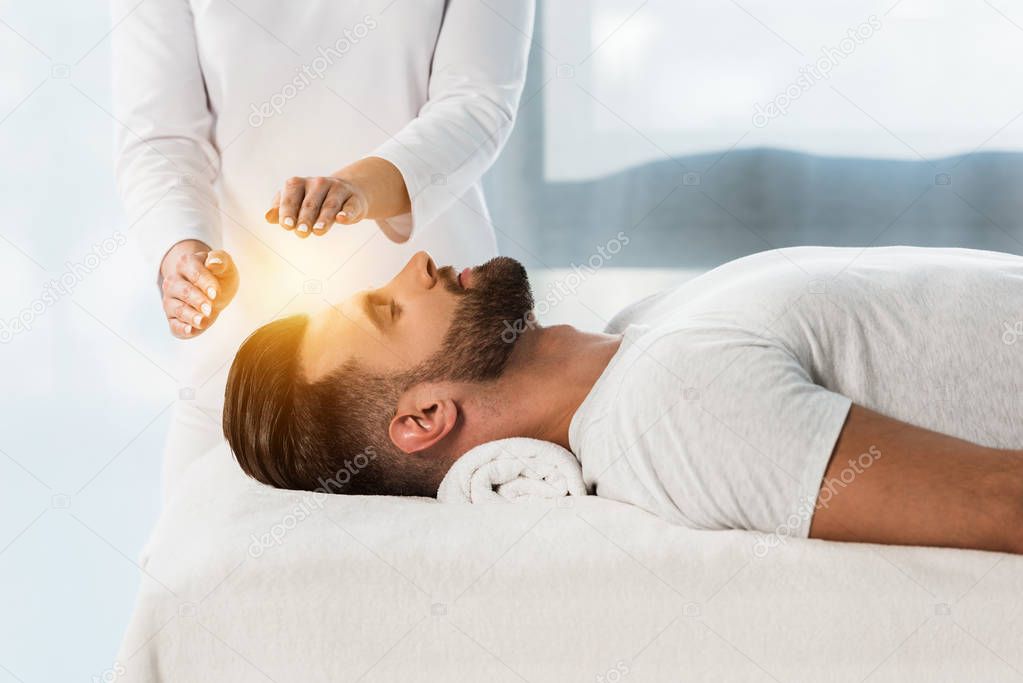 cropped view of healer putting hands above head of young man with closed eyes 