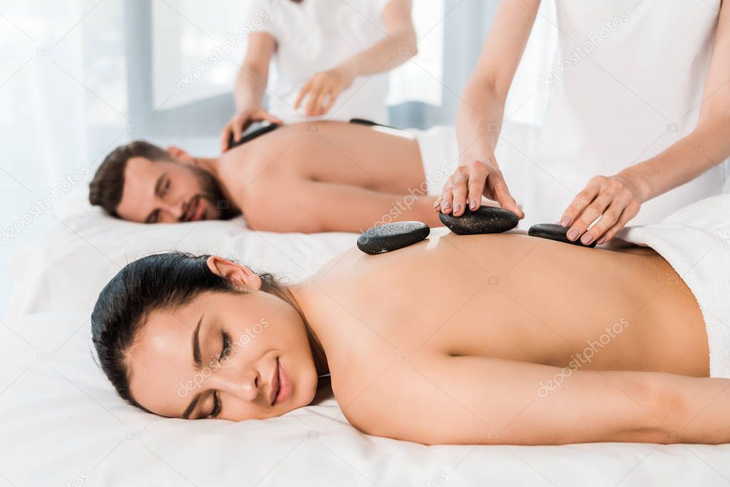 selective focus of masseur doing hot stone massage to attractive woman with closed eyes near bearded man 