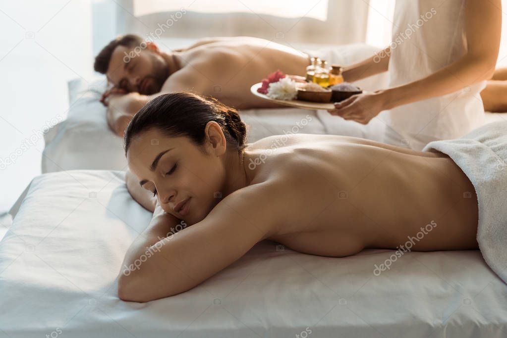 selective focus of masseur holding tray with sea salt and bottles of oil near woman and man in spa center 