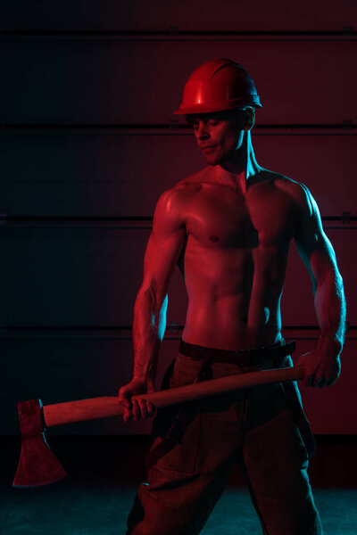 sexy shirtless fireman in protective helmet holding flat head axe in darkness
