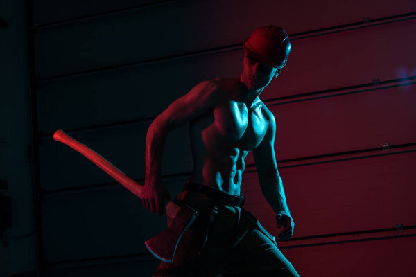 sexy shirtless fireman in protective hardhat holding flat head axe in darkness