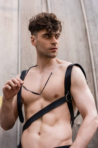 sexy shirtless man in sword belt holding sunglasses and looking away