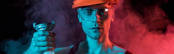 panoramic shot of serious fireman in hardhat and protective goggles holding fire hose in smoke on black