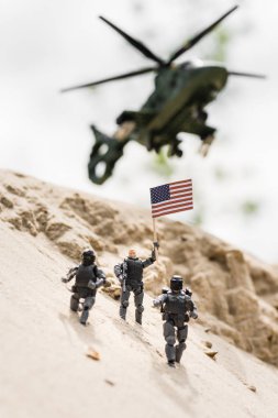 selective focus of toy soldier with weapon holding american flag on sand with helicopter on background clipart