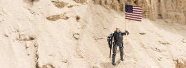 toy armed soldier standing on sand and holding american flag, panoramic shot clipart