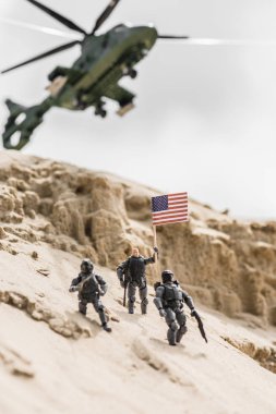 selective focus of toy soldiers on sand hill with american flag and helicopter on background clipart