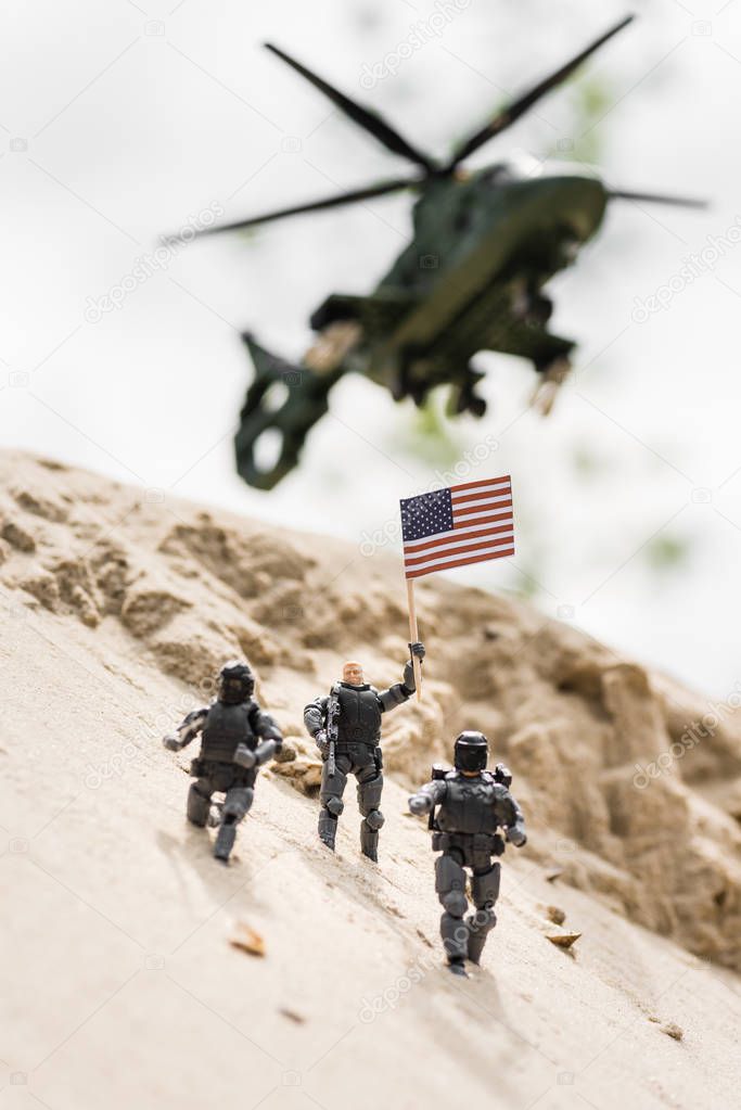 selective focus of toy soldier with weapon holding american flag on sand with helicopter on background