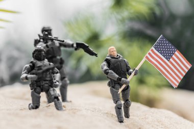 selective focus of toy soldiers aiming with guns at toy man with american flag on sand hill clipart