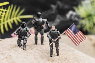 selective focus of toy soldiers with guns holding american flag on sand dune clipart