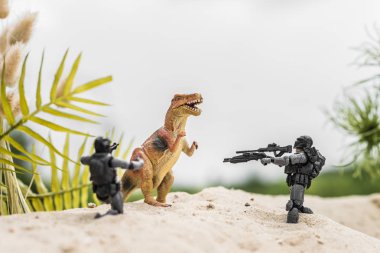 toy soldiers aiming with guns at toy dinosaur on sand hill clipart