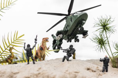 selective focus of plastic soldiers aiming at toy dinosaurs on sand dune with helicopter in sky clipart