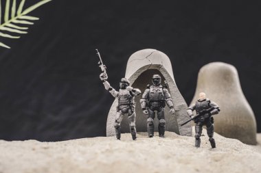 toy soldiers with guns and american flag standing near caves on sand dune on black background clipart