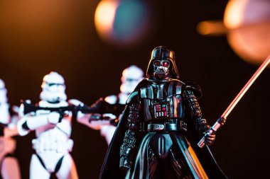 selective focus of Darth Vader with lightsaber and white imperial stormtroopers with guns on background clipart