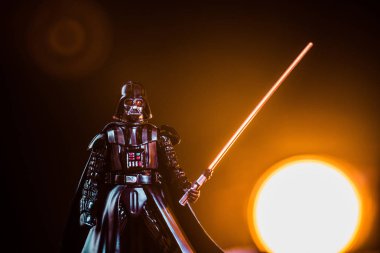 Darth Vader figurine with lightsaber on black background with shining sun clipart