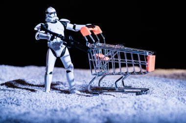 white plastic Imperial Stormtrooper with gun and shopping cart in space isolated on black