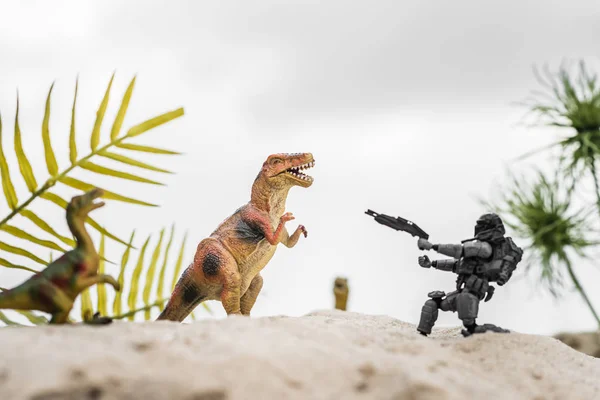 selective focus of plastic toy soldier aiming with gun at toy dinosaur on sand dune with tropical leaves