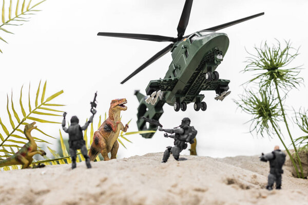 selective focus of plastic soldiers aiming at toy dinosaurs on sand dune with helicopter in sky