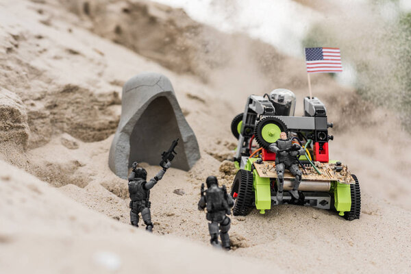 plastic toy soldiers on sand dune near car with american flag and cave