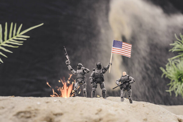 селективный фокус игрушечных солдат с оружием и американским флагом, стоящих рядом со взрывом на песчаной дюне
 