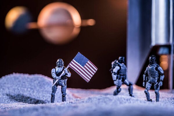 selective focus of toy astronauts holding american flag in space near rocket
