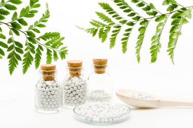 selective focus of small pills in glass bottles with wooden corks near spoon and green leaves on white 