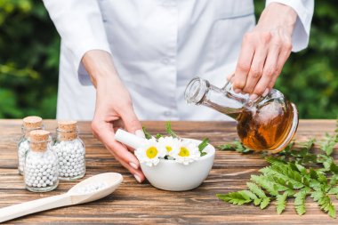 cropped view of woman holding jug with oil near mortar with flowers and green leaves on wooden table  clipart